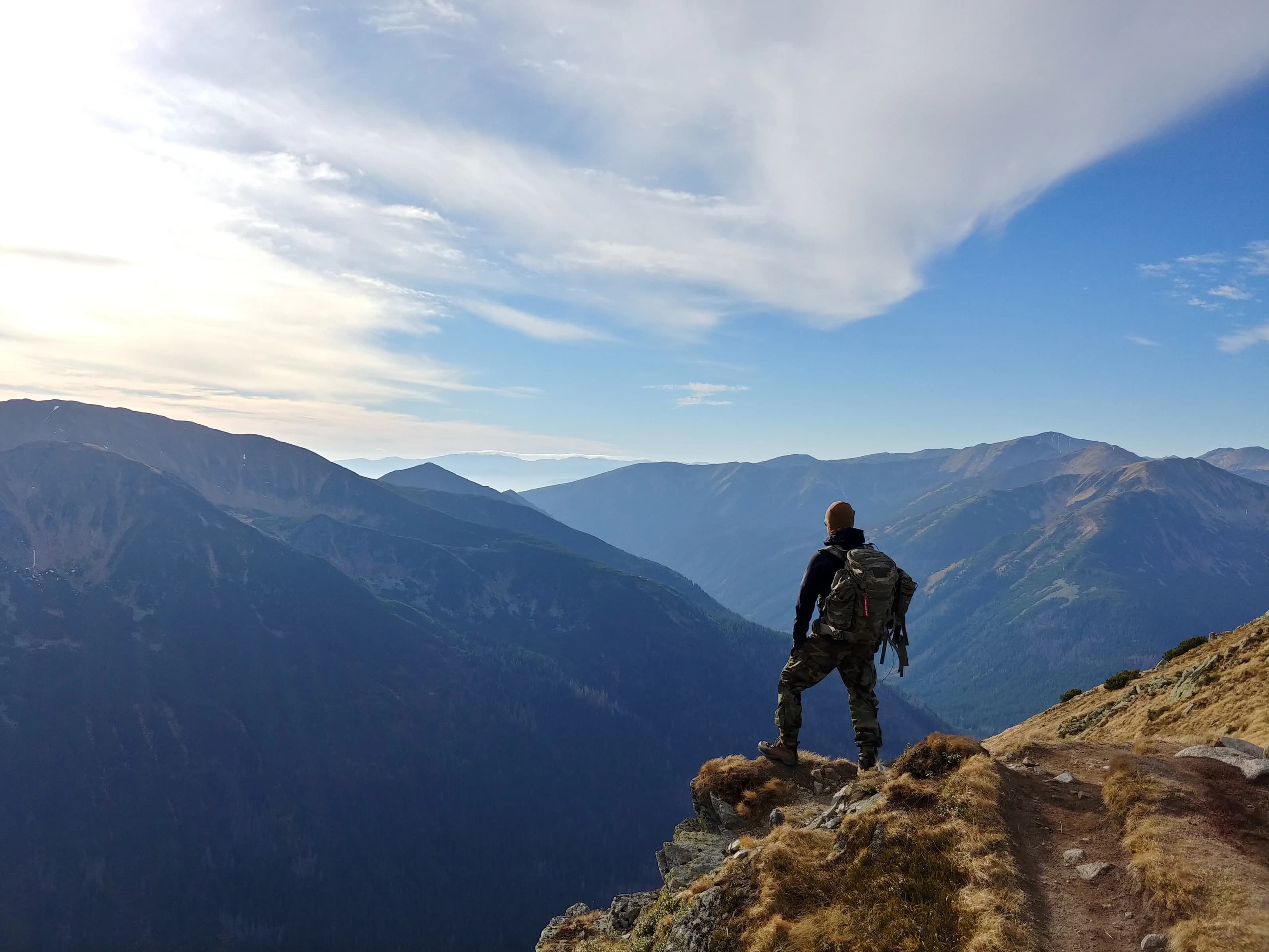 Hiker with a pack on a rocky ledge above a mountain valley under a clear blue sky
